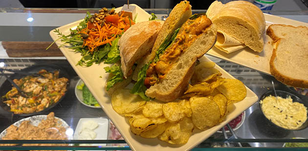 Image of a chicken chipotle sandwich on top of a deli counter. Through the see-through glass beneath, a range of sandwich fillings can be seen. On the plate is a sandwich surrounded by salad.
